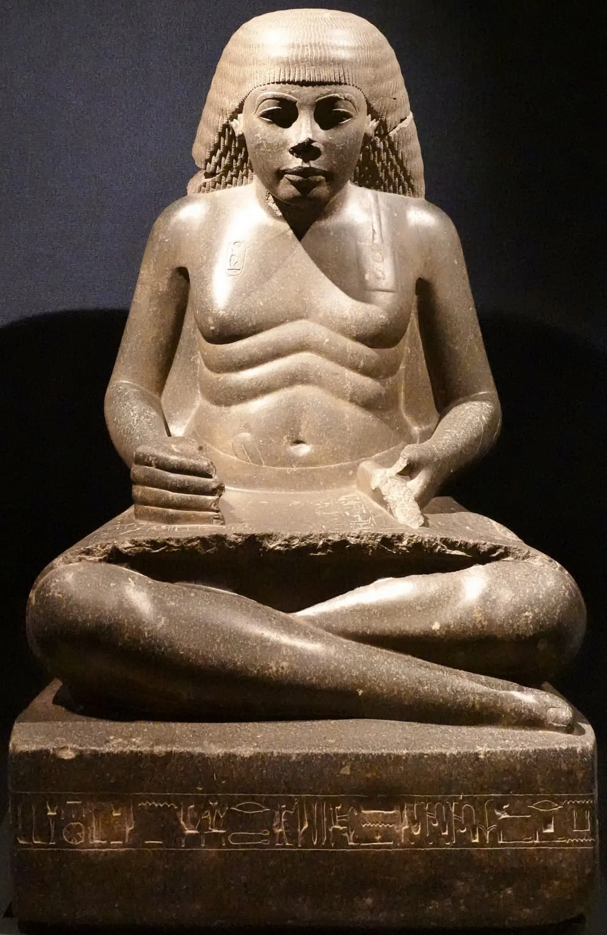 Sitzstatue aus dunklem Granit: ein Mann sitzt im Schneidersitz, trägt eine volle Perücke, deren gelockte Strähnen seitlich auf die Schultern fallen. Über den Bauch ziehen sich drei Hautfalten. Quer auf seinen Beinen liegt eine entrollte, mit Hieroglyphen beschriebene Papyrusrolle, an der Vorderkante abgebrochen. Eine Hand ruht auf dem Knie, die andere hält einen gerollten Gegenstand. Auf der Brust ist eine Königskartusche eingetieft, der Sockel trägt eine Zeile Hieroglyphen.