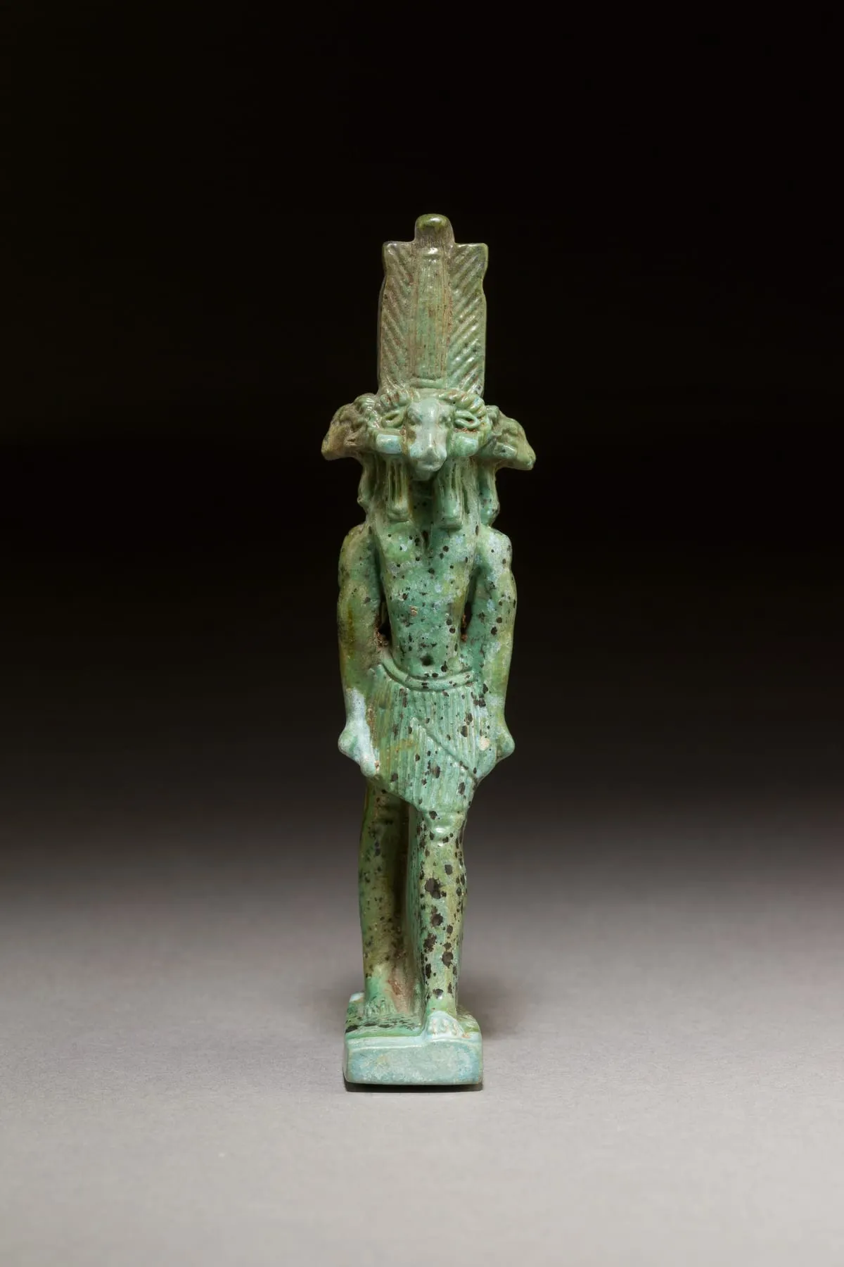 Kleine stehende Figur aus grün glasierter Fayence vor einem dunklen, nach unten heller werdenden Hintergrund. Der Körper ist menschlich, mit angelegten Armen und kurzem, senkrecht gefälteltem Schurz; die Beine stehen leicht versetzt auf einer rechteckigen Bodenplatte. Auf den Schultern sitzen mehrere Widderköpfe nebeneinander, deren Hörner waagerecht zur Seite gedreht sind — einer blickt nach vorn, weitere ragen nach links und rechts heraus. Darüber erhebt sich ein hoher Aufsatz aus zwei schmalen, längs gestreiften Federn mit einem runden Knauf dazwischen. Die Glasur ist ungleichmäßig und von zahlreichen dunklen Einsprengseln durchsetzt.