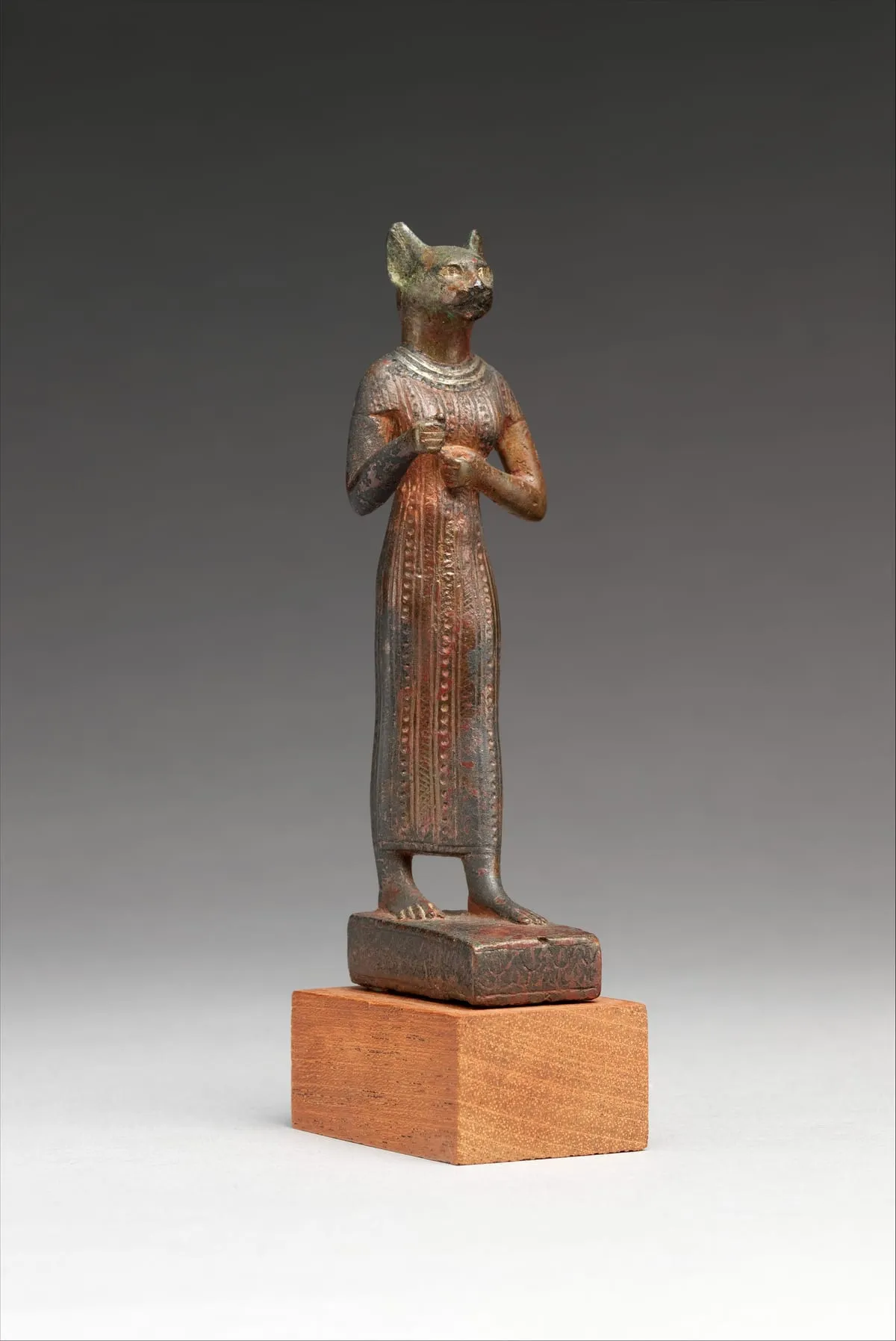 Stehende Frauenfigur aus Bronze mit rotbrauner Patina, auf einer flachen Standplatte. Der Kopf ist der einer Katze mit aufrecht stehenden spitzen Ohren; die Augen sind mit hellem Metall eingelegt und heben sich glänzend ab. Sie trägt ein enges, knöchellanges Kleid mit feinen senkrechten Rippen und Reihen von Punktverzierung, dazu einen Halskragen. Beide Arme sind angewinkelt, die Hände halten vor der Brust einen kleinen Gegenstand.