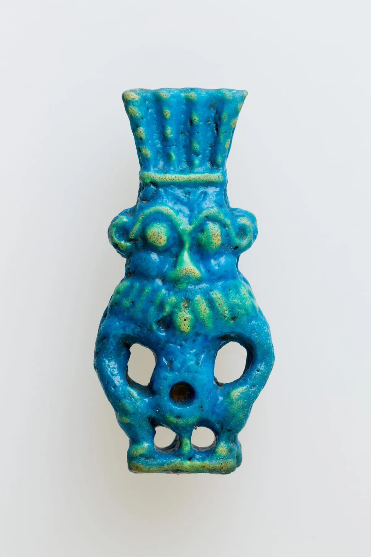 Kleines Amulett aus leuchtend türkisblauer Fayence, frontal dargestellt: ein untersetzter Zwerg mit breitem, flachem Gesicht, großen runden Augen und breiter Nase. Um das Gesicht liegt ein gerippter Kranz aus Mähne und Bart. Auf dem Kopf sitzt über einem Querband eine hohe, nach oben ausschwingende Federkrone. Die kurzen Arme sind angewinkelt, zwischen Armen und Rumpf sowie zwischen den gebogenen Beinen ist der Ton durchbrochen. In der Bauchmitte sitzt eine runde Vertiefung.