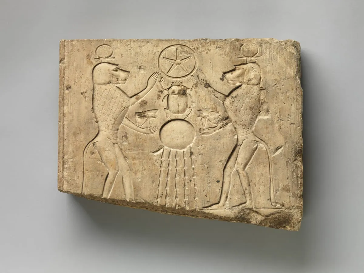 Querformatige Kalksteinplatte mit flachem Relief. In der Mitte ein großer Käfer von oben gesehen; über ihm eine Scheibe mit sternförmigem Muster, unter ihm eine zweite Scheibe, von der senkrechte Strahlen nach unten fallen. Links und rechts steht je ein aufrecht hockender Pavian im Profil, dem Käfer zugewandt, eine Hand erhoben, in der anderen ein Udjat-Auge. Beide tragen eine Scheibe auf dem Kopf.
