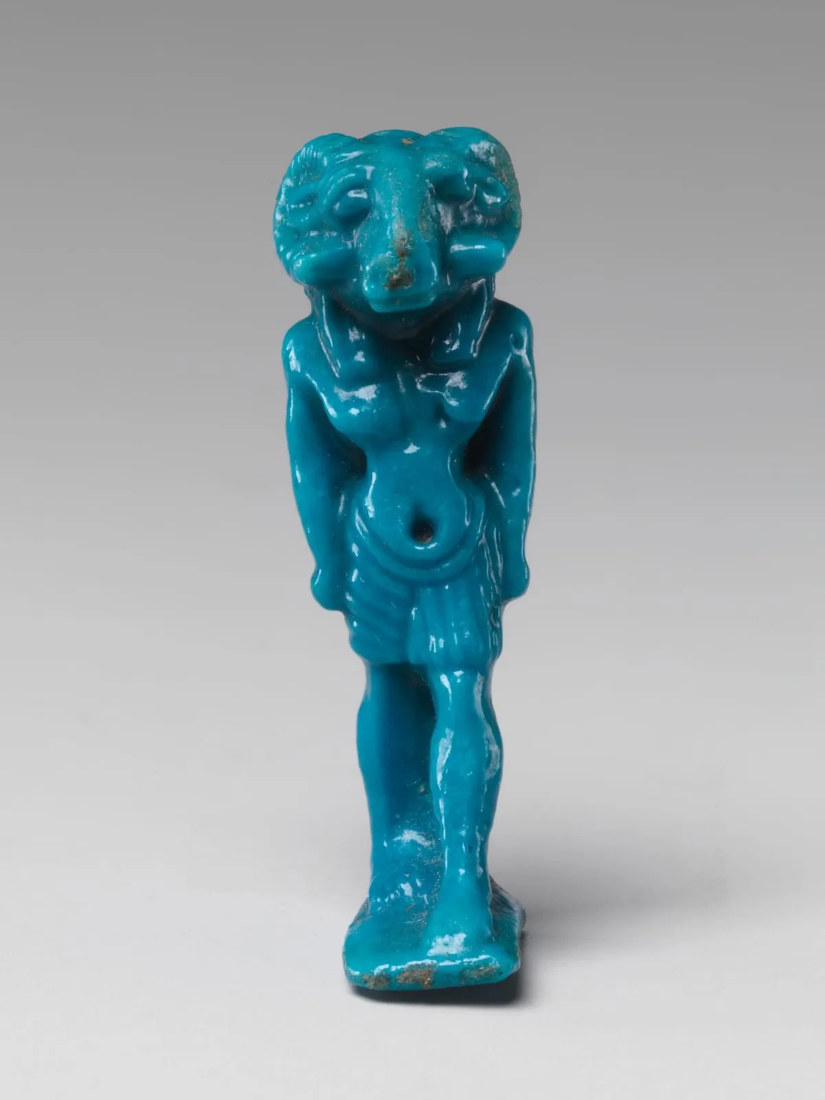 Stehende Amulettfigur aus türkisfarbener Fayence: ein Mann mit dem Kopf eines Widders. Die Hörner schwingen waagerecht zu beiden Seiten aus dem Kopf heraus, die Schnauze ist breit. Der Oberkörper ist nackt, um die Hüften liegt ein gefältelter Schurz. Die Arme hängen seitlich herab, die Figur steht auf einer angearbeiteten Platte. In den Vertiefungen hat sich die Glasur dunkel abgesetzt.