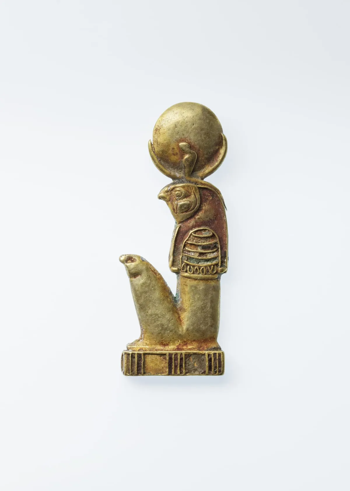 Kleiner Anhänger aus Gold, die Figur im Profil nach links. Ein kniender Körper mit Falkenkopf, dessen Auge und Schnabel deutlich ausgearbeitet sind. Auf dem Kopf ruht eine große runde Scheibe, die von einer nach oben geöffneten Sichel getragen wird; vorn an der Scheibe sitzt eine kleine aufgerichtete Schlange. Über der Brust liegt ein breiter Kragen in mehreren Reihen. Die Figur steht auf einem gerippten Sockel.