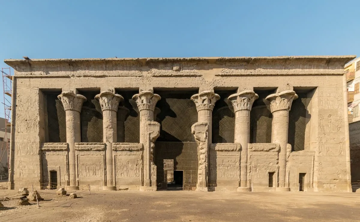 Frontalansicht einer breiten, rechteckigen Tempelfassade aus graubraunem Sandstein. Sechs kräftige Säulen mit unterschiedlich gestalteten Pflanzenkapitellen stehen in einer Reihe; zwischen ihnen sitzen brusthohe Schrankenmauern, in der Mitte ein vergitterter Eingang. Alle Flächen sind mit flachen Reliefbändern und Figurenreihen bedeckt, oben schließt ein glatter Architrav ab. Der Boden davor liegt tiefer als die Umgebung. Am rechten Bildrand ragt ein modernes Wohnhaus mit Fenstern und Balkonen unmittelbar an die Tempelwand heran, links steht ein Baugerüst.