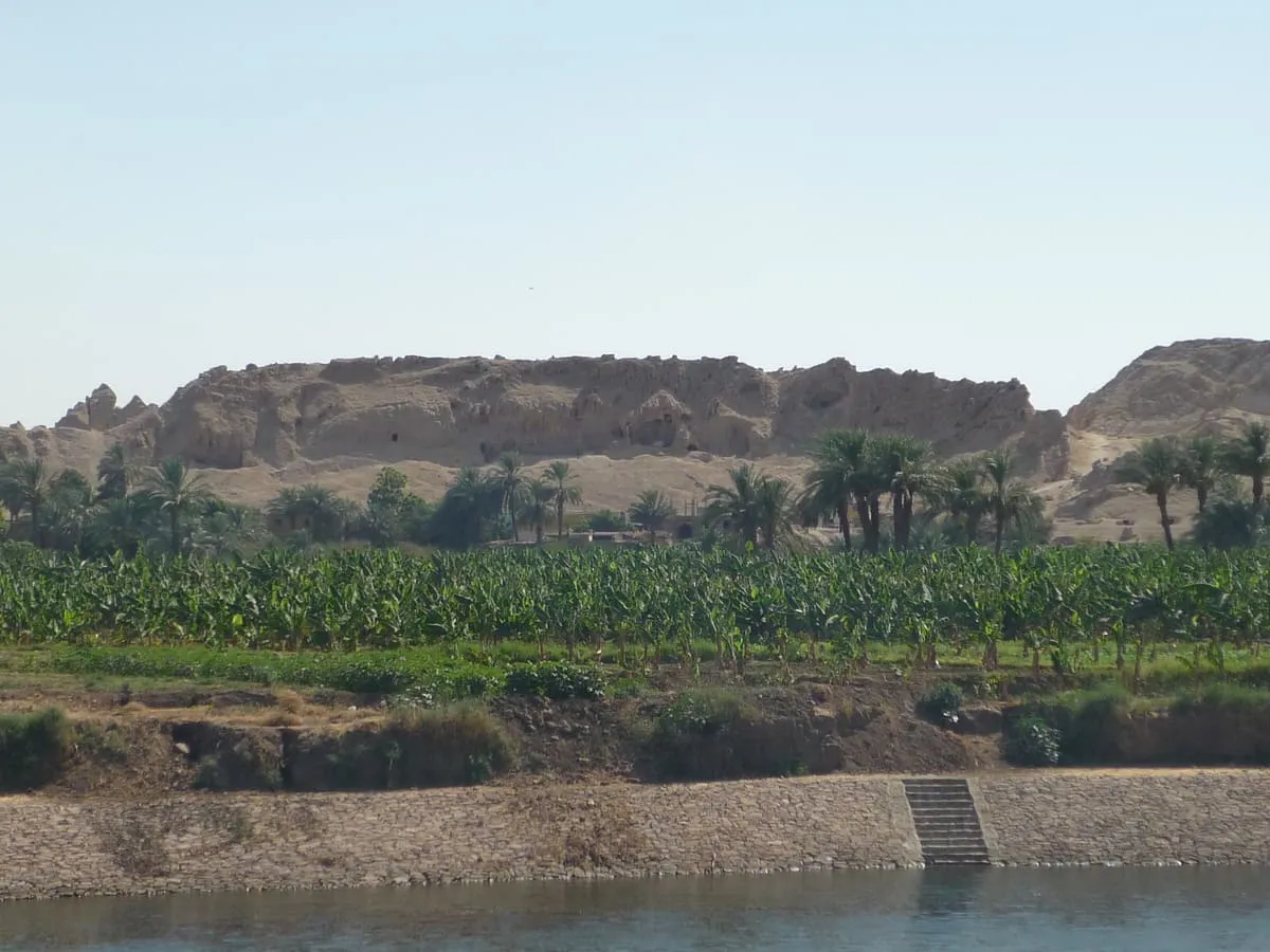 Blick vom Nil auf einen langgestreckten, hellbraunen Felshügel. Im Vordergrund das Wasser und eine gepflasterte Uferböschung mit einer schmalen Treppe; darüber ein grüner Streifen aus Bananenstauden und Feldern, dahinter eine Reihe Dattelpalmen und einzelne Häuser. Die Hügelflanke ist stark verwittert und zeigt mehrere dunkle Höhlungen und Einschnitte; rechts schließt eine zweite Erhebung an.