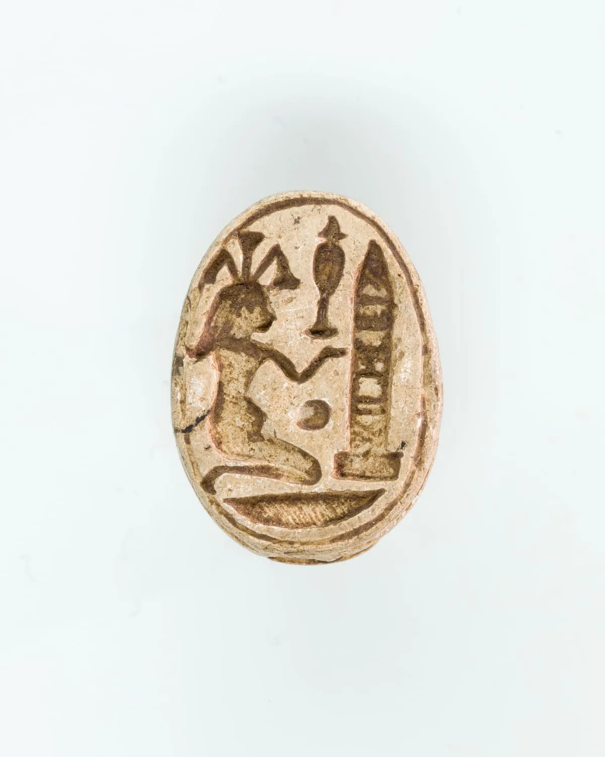 Unterseite eines hellen Steinskarabäus, oval gerahmt. Eingeschnitten kniet eine Gestalt im Profil nach rechts, aus deren Kopf mehrere Pflanzenstengel fächerförmig nach oben wachsen. Ein Arm ist nach vorn gestreckt. Davor stehen ein hohes schmales Gefäß und ein aufrechter, innen gegliederter Pfeiler; darunter liegt ein waagerechtes Zeichen.