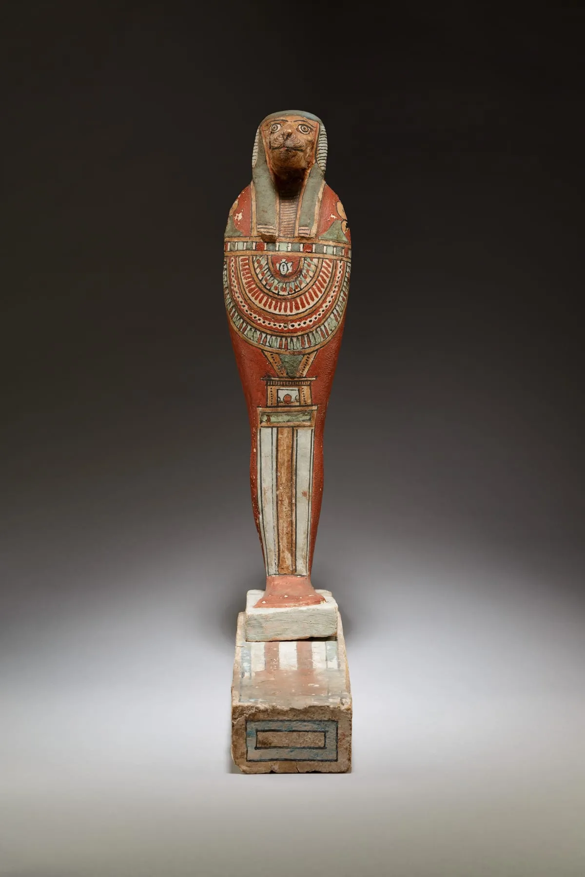 Bemalte Holzfigur in Mumienform auf einem gestuften Sockel. Der Kopf ist der eines Pavians mit vorspringender Schnauze, gerahmt von einem gestreiften Kopftuch, das auf die Schultern fällt. Über der Brust liegt ein breiter Halskragen aus vielen farbigen Bögen in Rot, Weiß und Grün. Darunter zieht sich ein senkrechtes Schriftfeld bis zu den Füßen. Der Körper ist rotbraun gefasst.