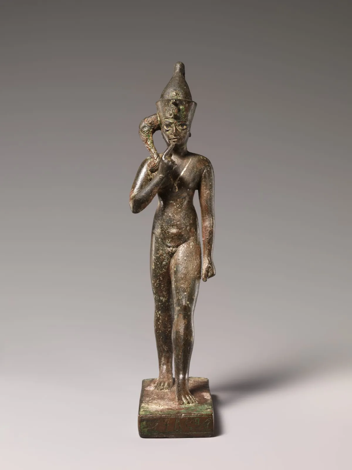 Stehende Bronzefigur eines nackten Knaben auf beschrifteter Basis. Ein Arm ist angewinkelt erhoben, der Zeigefinger berührt den Mund. Der andere Arm hängt herab. Von der Schläfe fällt eine dicke, geflochtene Locke über die Schulter. Auf dem Kopf sitzt eine hohe kegelförmige Krone mit einer kleinen aufgerichteten Schlange an der Stirn. Ein Bein ist leicht vorgesetzt.