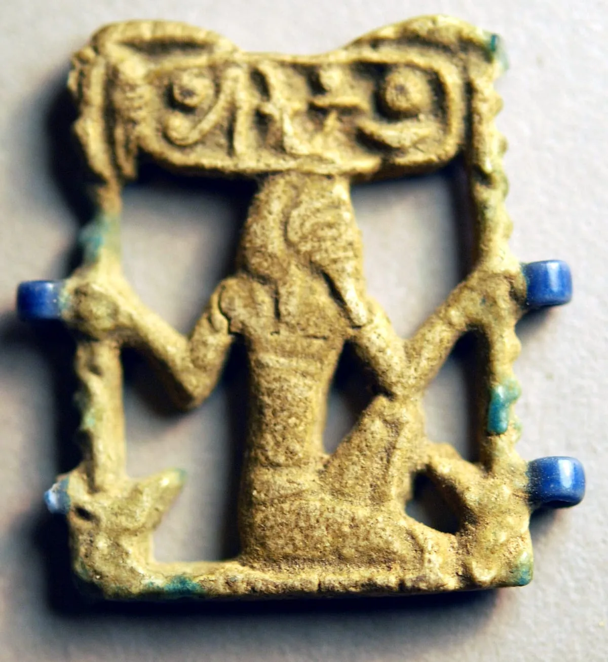 Kleines durchbrochenes Amulett aus gelblicher Fayence, von oben auf hellem Grund aufgenommen. In der Mitte kniet eine Gestalt: Der Oberkörper ist frontal gegeben, der Kopf im Profil, die Arme sind waagerecht zu beiden Seiten ausgestreckt. Von jeder Hand läuft ein schmaler, quer gekerbter Streifen schräg nach unten zum Rand des Amuletts. Über der Gestalt liegt ein waagerechter Balken mit einer von einem Oval umschlossenen Zeichengruppe; unten schließt eine gerade Leiste ab. An drei Stellen ragen kurze, kräftig blaue Stifte aus dem Rand. Die Oberfläche ist abgerieben, in den Vertiefungen haben sich türkisfarbene Glasurreste erhalten.