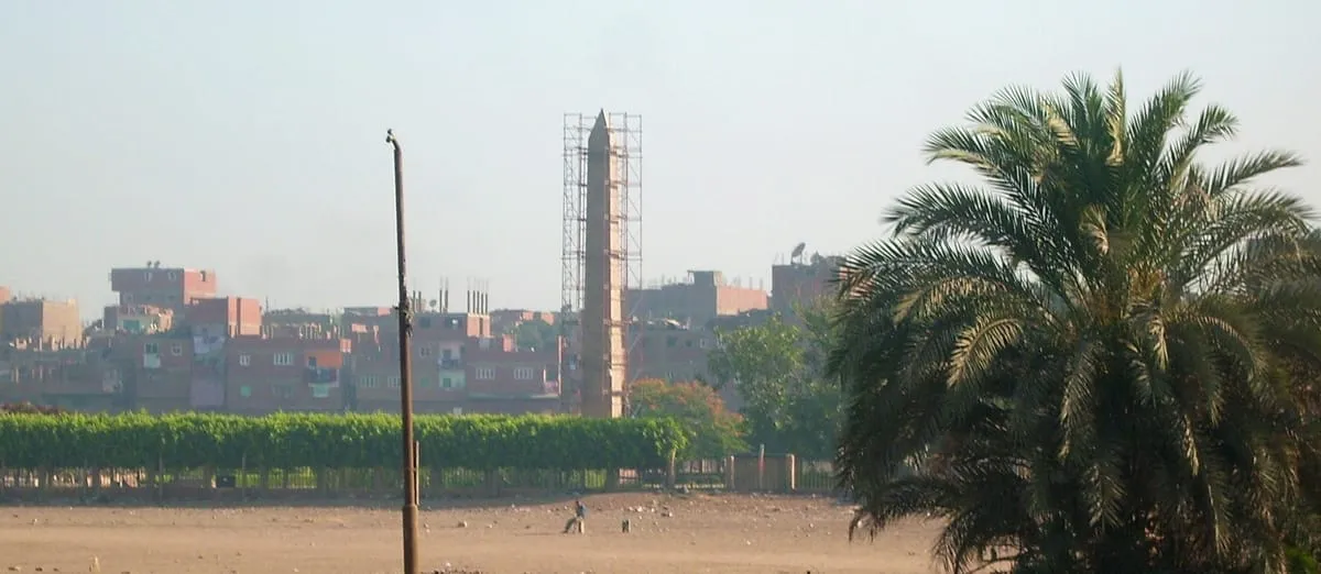 Weiter Blick über ein staubiges, offenes Gelände auf eine Stadt im Dunst. In der Bildmitte ragt ein schlanker steinerner Obelisk mit pyramidenförmiger Spitze auf; er ist bis knapp unter die Spitze von einem Gerüst aus dünnen Stangen umgeben. Ringsum stehen dicht gedrängt mehrstöckige Wohnhäuser aus rotem Backstein mit flachen Dächern, dazwischen ein Streifen dunkelgrüner Bäume und eine Mauer. Links steht ein Strommast, rechts füllt eine große Dattelpalme den Bildrand. Im Vordergrund geht eine einzelne kleine Gestalt über den Sand.