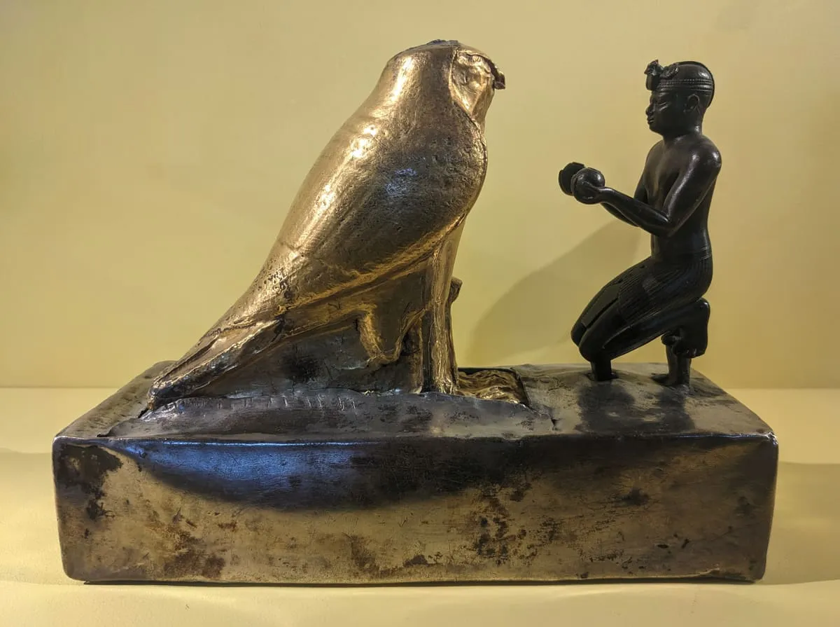 Kleine Figurengruppe auf einem massiven, silbrig glänzenden Sockel vor hellem Grund. Links steht ein großer, vollständig vergoldeter Falke im Profil, aufrecht, mit angelegten Flügeln und deutlich gezeichnetem Gefieder; die Oberseite seines Kopfes ist abgebrochen. Rechts kniet ihm zugewandt eine kleinere dunkle Metallfigur: ein Mann mit eng anliegender Kopfbedeckung und gefälteltem Schurz, der mit beiden Händen ein rundes Gefäß vor die Brust hält. Der Falke überragt die kniende Gestalt deutlich.