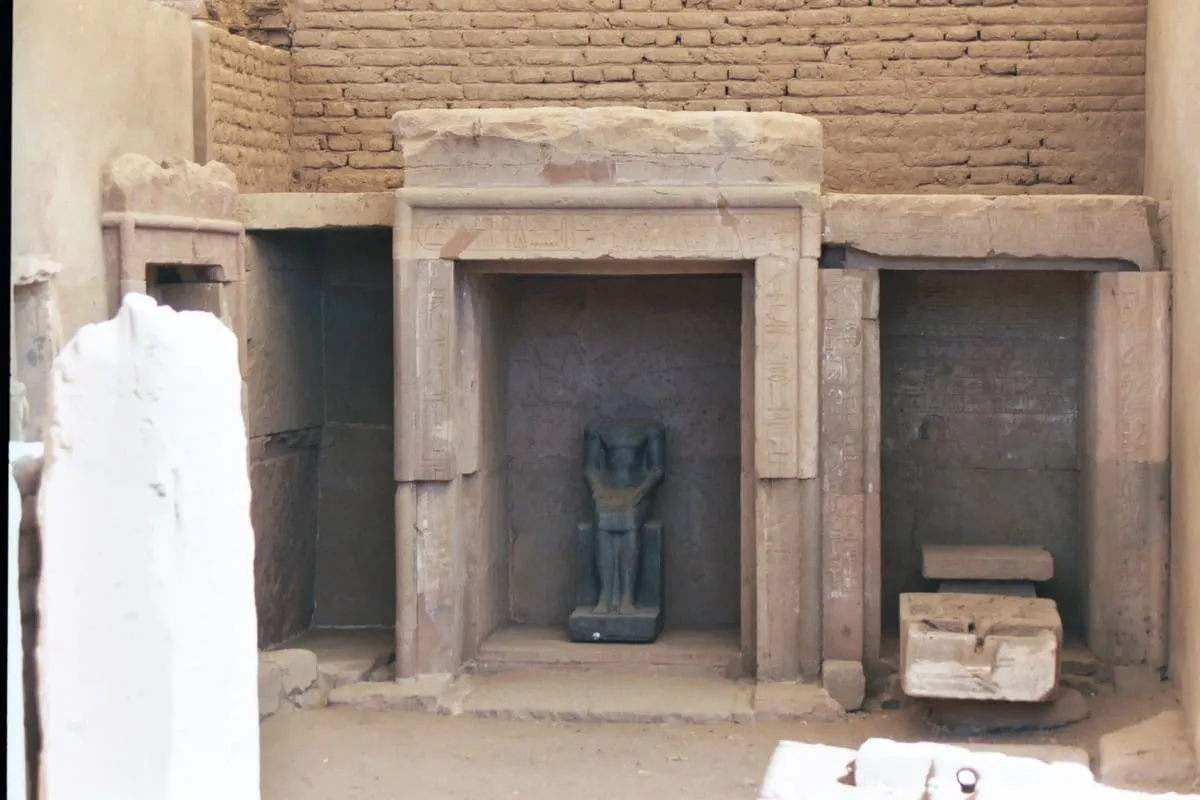 Ausgrabungsbefund im Freien, quer aufgenommen. Vor einer hohen Wand aus luftgetrockneten Lehmziegeln stehen nebeneinander drei aus Stein gefügte Nischen; ihre Pfosten und Türstürze sind mit Hieroglyphen beschriftet. Die mittlere und größte Nische ist offen und enthält eine dunkle Steinfigur auf einem niedrigen Sockel: ein Mensch in aufrechter Haltung, die Arme am Körper, das Gesicht stark verwittert. Die linke Nische ist leer und verschattet, in der rechten steht eine unbeschriftete Platte. Davor liegen auf sandigem Boden mehrere Steinblöcke und Bruchstücke, links am Bildrand ein hell verputzter Pfeiler. Die Farben sind gedämpft, die Aufnahme leicht körnig.