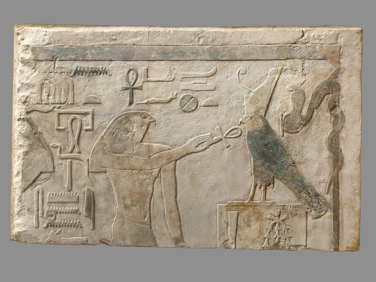 Querformatiger Kalksteinbalken mit erhabenem Relief, Farbreste in Blaugrün. Links steht ein Mann im Profil mit dem Kopf eines Falken; sein Arm reicht nach rechts und hält ein Anch-Zeichen. Rechts sitzt ein Falke auf einem hohen rechteckigen Sockel, in dessen Feld Zeichen stehen; Gefieder und Flügel sind noch bläulich gefärbt. Darüber und dazwischen stehen einzelne Hieroglyphen, am rechten Rand ragt eine aufgerichtete Schlange auf. Am oberen Rand verläuft ein farbiges Band.