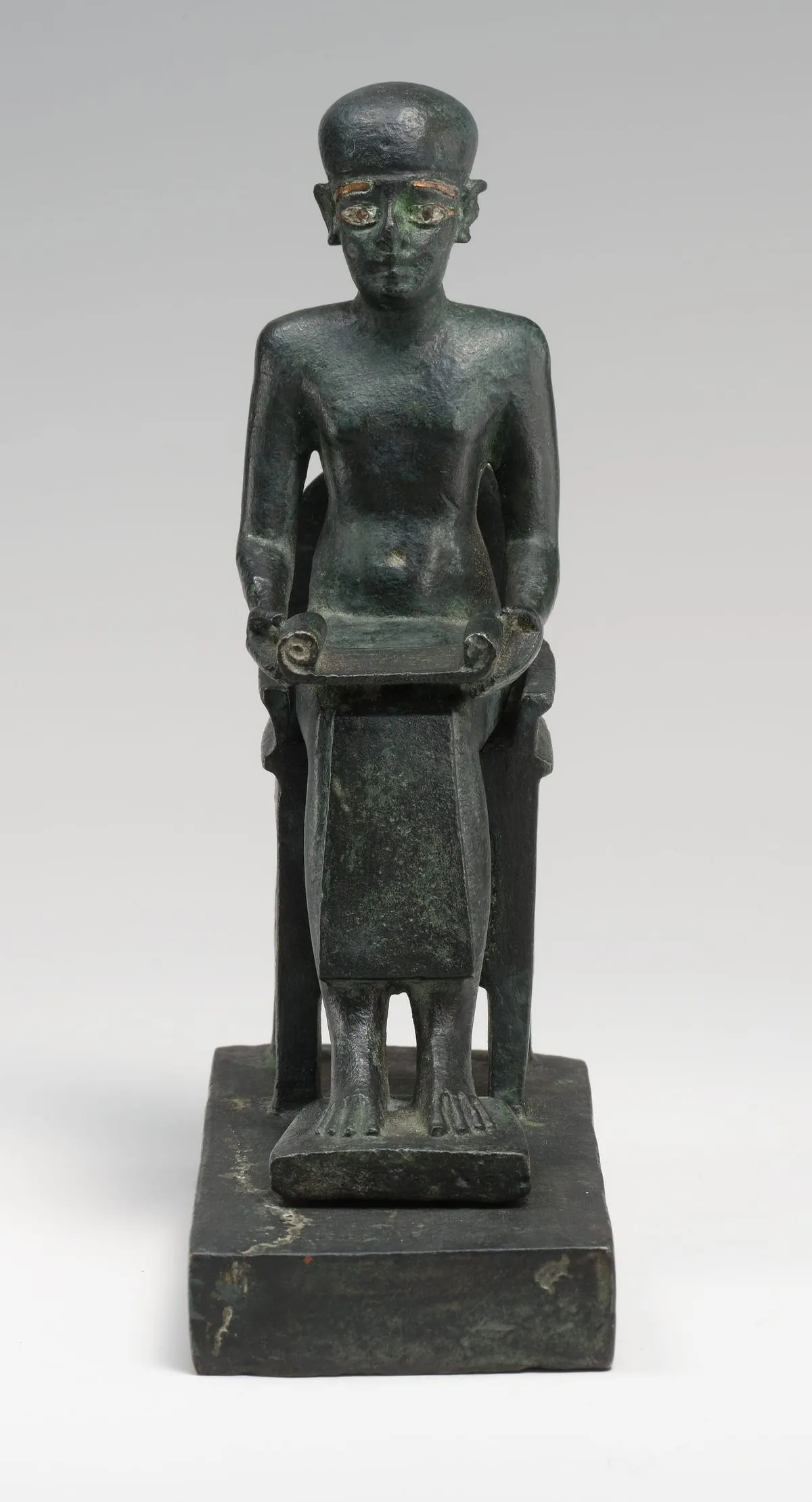 Kleine sitzende Bronzefigur vor hellgrauem Hintergrund, frontal aufgenommen. Der Mann trägt eine glatte, eng anliegende Kappe, die die Ohren frei lässt; Augenbrauen und Augenränder sind mit einem helleren Metall eingelegt, die Augäpfel hell abgesetzt. Der Oberkörper ist unbekleidet und schlicht modelliert. Über den Oberschenkeln liegt waagerecht eine ausgerollte Schriftrolle, deren beide Enden zu Spiralen eingedreht sind; die Hände fassen sie an den Seiten. Darunter fällt ein glatter Schurz bis über die Knie. Die Füße stehen nebeneinander auf einer schmalen Platte, die Zehen sind einzeln ausgearbeitet; die Figur sitzt auf einem kastenartigen Stuhl ohne Lehne und steht auf einem massiven, dunklen Sockel. Die Oberfläche ist dunkelgrün patiniert und stellenweise heller ausgewaschen.