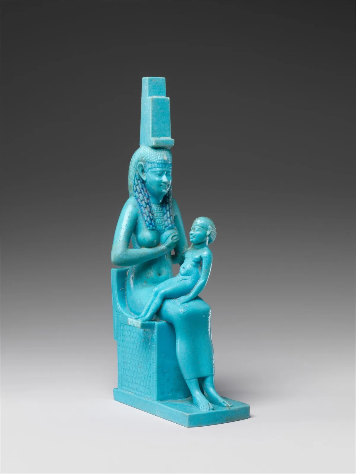 Türkisfarbene Fayencestatuette: eine Frau sitzt auf einem Blockthron, trägt eine lange Strähnenperücke und auf dem Kopf das Thronzeichen. Quer auf ihrem Schoß sitzt ein nacktes Kind, das sie stillt.