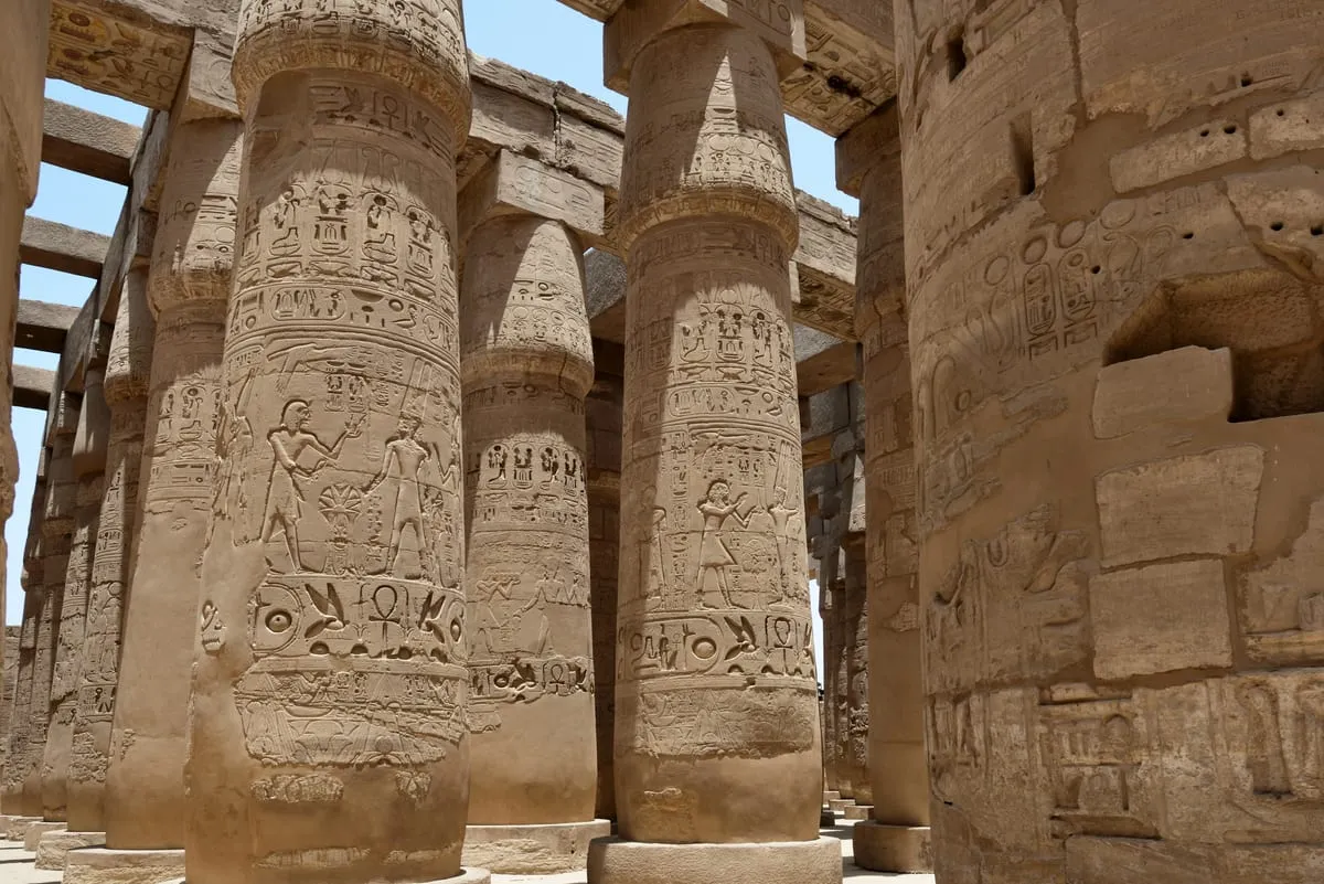 Blick von unten zwischen mächtige Steinsäulen eines offenen Tempelsaals. Die Säulen stehen dicht in mehreren Reihen; ihre Schäfte sind über die ganze Höhe mit flach eingetieften Reliefs und Hieroglyphenbändern bedeckt, darunter Szenen mit stehenden Figuren, die einander gegenüberstehen. Oben liegen schwere Steinbalken quer auf den Kapitellen; dazwischen fällt Licht aus einem klaren blauen Himmel ein. Rechts im Bild schließt eine hohe, ebenfalls beschriftete Wand an, aus der einzelne Blöcke herausgebrochen sind. Der Stein ist warm ockerfarben, das Licht hart, die Schatten kurz.