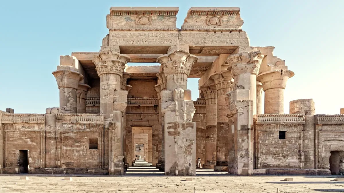 Frontalansicht einer Tempelfassade aus hellem Sandstein, streng symmetrisch aufgebaut. In der Mitte steht ein breiter Pfeiler; links und rechts davon öffnet sich je ein gleich großer Durchgang, flankiert von Säulen mit Pflanzenkapitellen. Über den beiden Öffnungen liegt je ein Architrav mit einer geflügelten Sonnenscheibe. Vorn schließen brusthohe Schrankenmauern mit Reliefs an. Durch die linke Öffnung sieht man in eine Flucht weiterer Tore; eine kleine Figur gibt den Maßstab. Der Himmel ist wolkenlos.