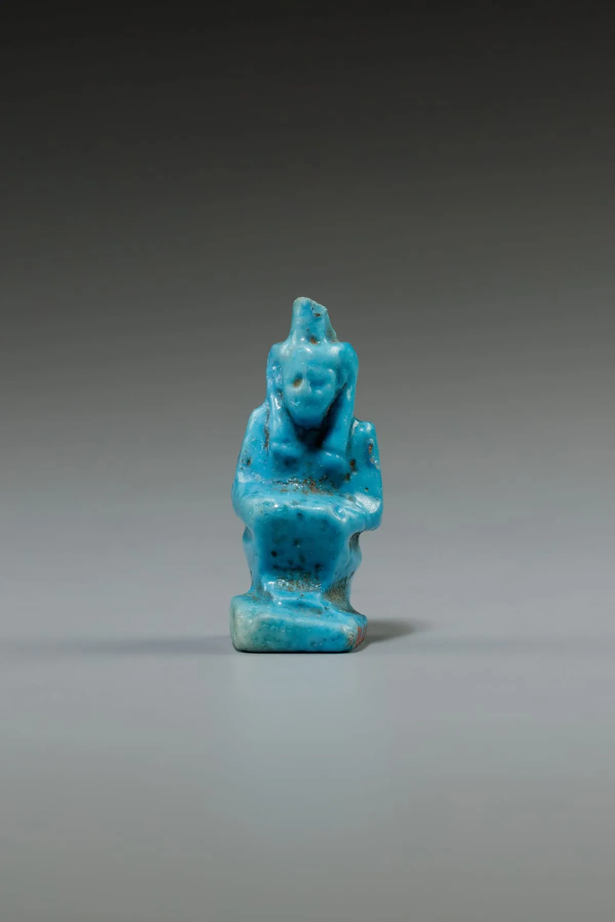 Kleine Amulettfigur aus leuchtend blauer Fayence: eine Gestalt hockt mit angezogenen Knien auf einer flachen Platte, die Hände liegen auf den Knien. Vom Kopf steht ein schmales, nach hinten geneigtes Blatt auf. Die Oberfläche ist abgerieben, die Gesichtszüge sind kaum noch zu erkennen.