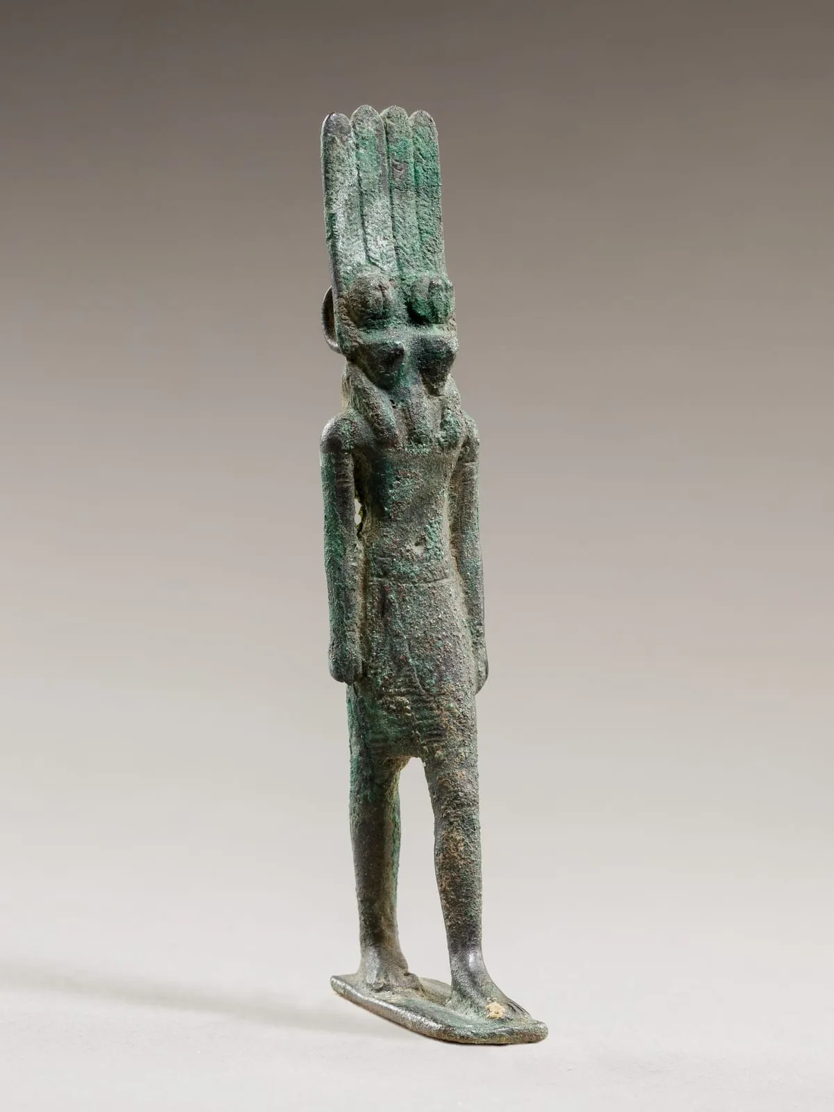 Kleine Bronzefigur mit grüner Patina auf einer Standplatte. Der Körper ist der eines schreitenden Mannes mit nacktem Oberkörper und kurzem Schurz, die Arme hängen seitlich herab. Auf den Schultern sitzen zwei Falkenköpfe nebeneinander, jeder mit Schnabel und rundem Auge. Darüber erhebt sich eine hohe Krone aus mehreren schmalen, senkrecht gerippten Federn.
