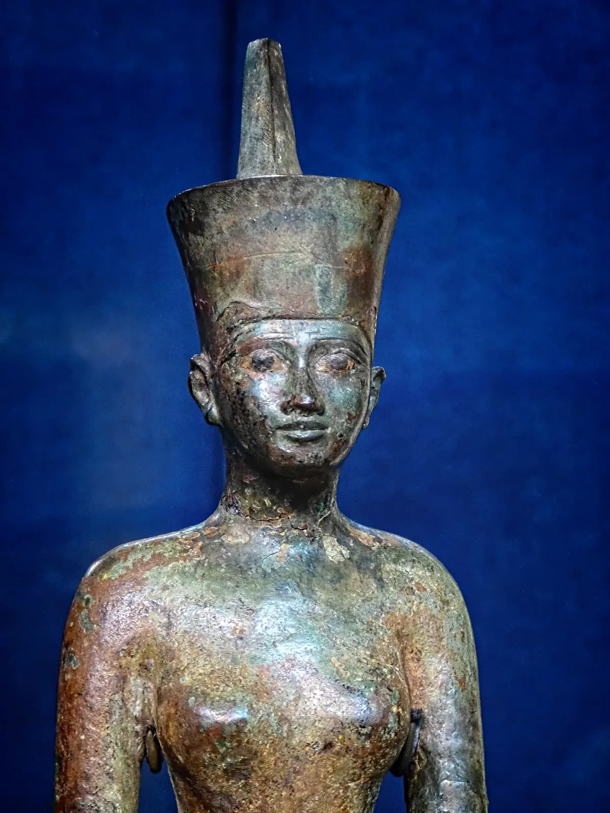 Oberkörper einer Bronzefigur vor dunkelblauem Grund, frontal aufgenommen und unterhalb der Brust angeschnitten. Auf dem Kopf sitzt eine hohe Kopfbedeckung: ein nach oben leicht ausgestellter zylindrischer Teil, aus dem hinten ein schmaler, spitz zulaufender Schaft weit aufragt. Unter dem Rand verläuft ein schmales Band über die Stirn. Das Gesicht ist breit und ruhig, mit mandelförmigen Augen unter erhabenen Lidrändern, gerader Nase und geschlossenen Lippen; die Ohren stehen groß und weit vom Kopf ab. Am Hals liegen mehrere waagerechte Ringe. Die Schultern sind unbekleidet, die Brüste ausgearbeitet. Die Oberfläche ist fleckig grün, braun und rötlich korrodiert, an Wangen, Schlüsselbeinen und Brust bis zu einem hellen, metallischen Schimmer abgerieben.