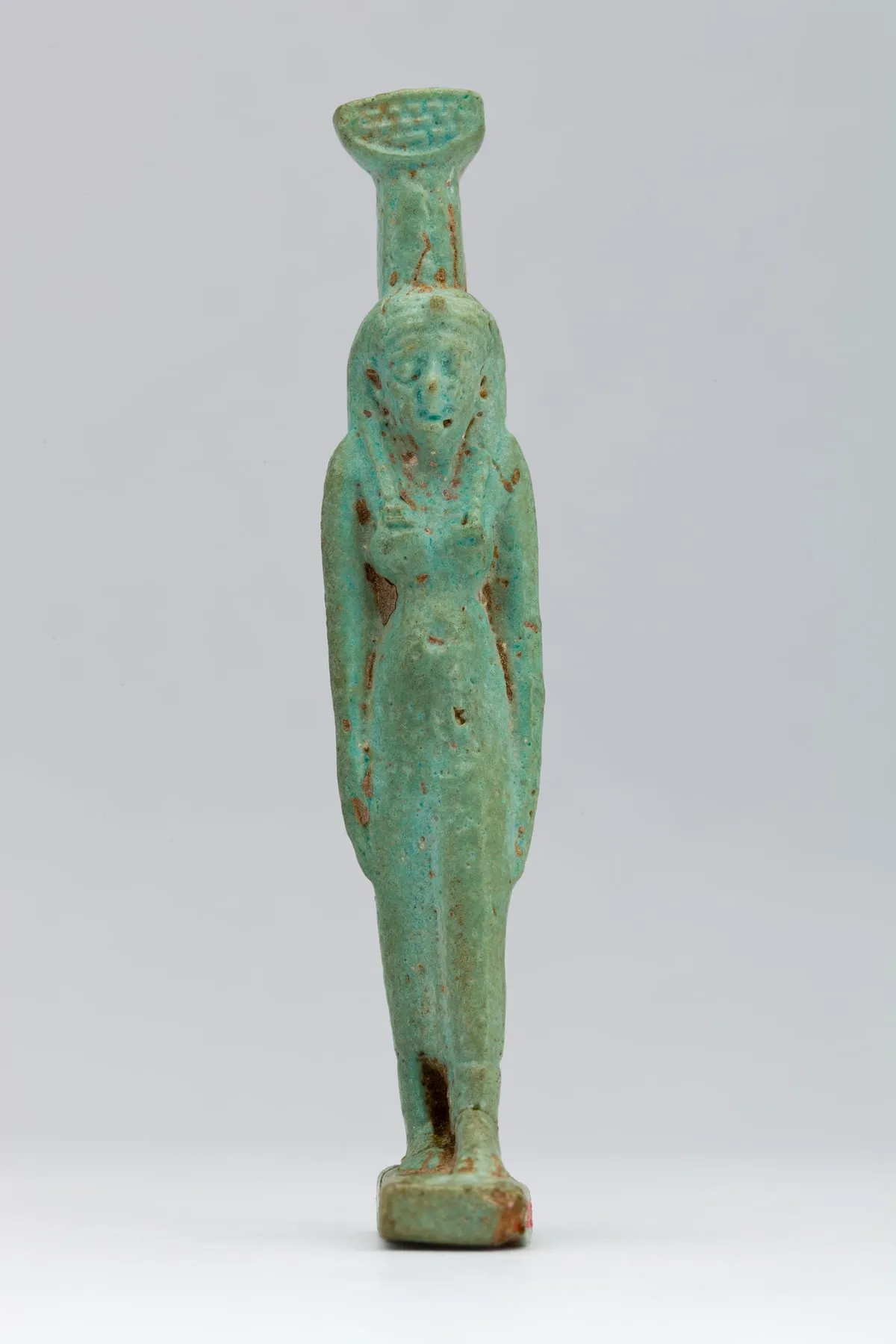 Stehende Amulettfigur aus grüntürkiser Fayence auf einer kleinen Platte. Die Frau trägt eine lange Strähnenperücke, die beidseits auf die Schultern fällt, und ein enges knöchellanges Kleid; die Arme liegen dicht am Körper. Auf dem Kopf steht ein schmaler aufrechter Pfeiler, darüber ein breiter, flach ausschwingender Korb mit geflochtenem Muster. An abgeplatzten Stellen tritt der rötliche Scherben hervor.