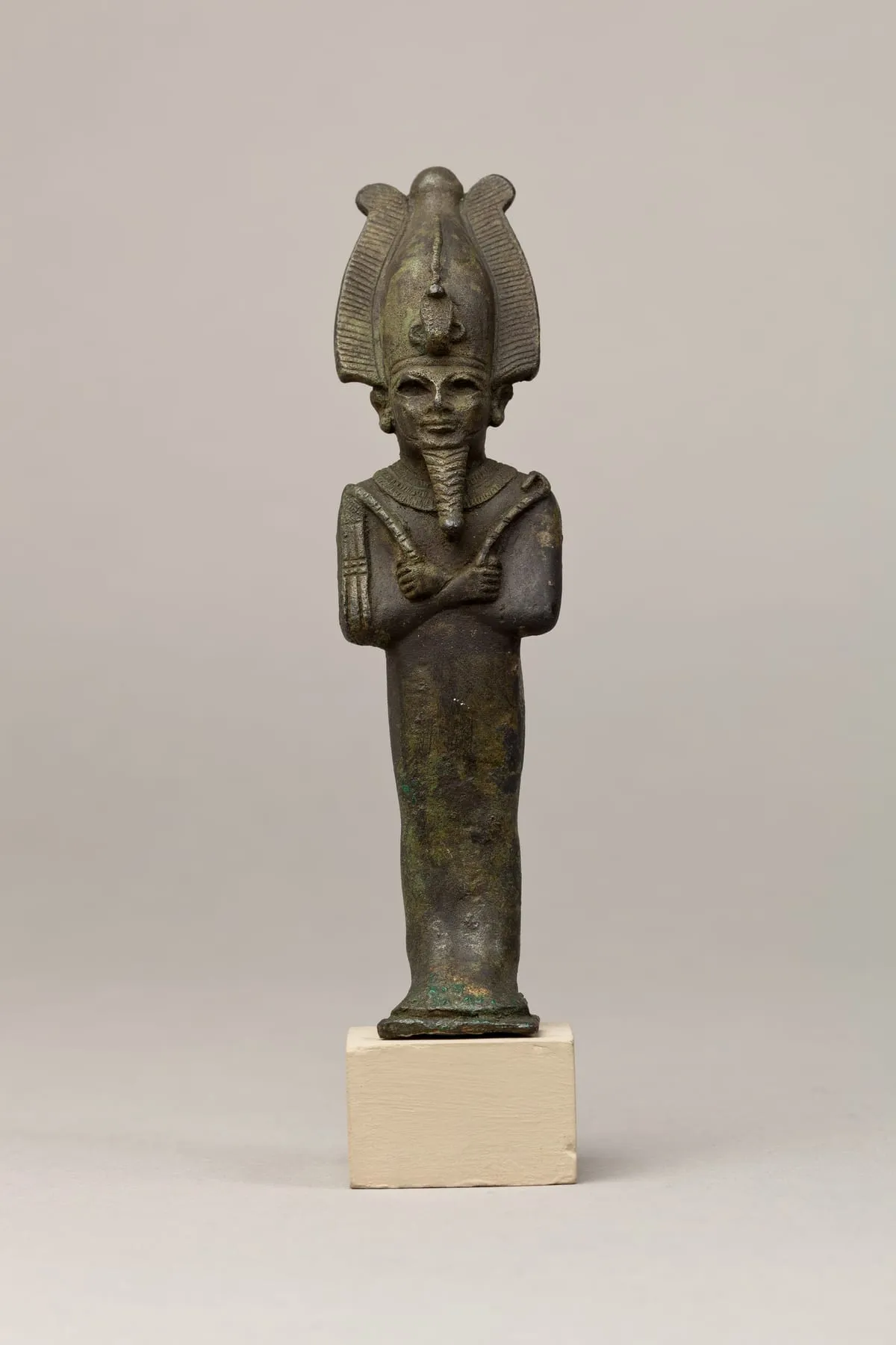 Stehende Bronzefigur in Mumienform auf einer Standplatte. Der Körper ist glatt umwickelt und verjüngt sich nach unten. Beide Arme liegen gekreuzt vor der Brust; die Hände treten aus der Umwicklung und halten einen Krummstab und einen Wedel. Vom Kinn hängt ein langer geflochtener Bart. Auf dem Kopf sitzt eine hohe Krone: ein Mittelkegel mit kleinem Knauf, flankiert von zwei großen gerippten Federn, vorn eine kleine aufgerichtete Schlange.