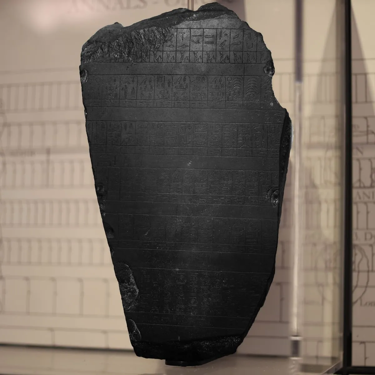 Eine dunkle, annähernd trapezförmige Steinplatte steht aufrecht in einer Vitrine. Ihre Oberfläche ist von waagerechten Linien in ein Dutzend schmaler Register geteilt, die dicht mit eingetieften Hieroglyphen gefüllt sind; senkrechte Striche unterteilen die Register zusätzlich in kurze Fächer. Der obere Rand ist unregelmäßig abgebrochen, an den Seiten fehlen Stücke. Im Hintergrund hängt eine helle Tafel mit einer gezeichneten Umschrift derselben Register.