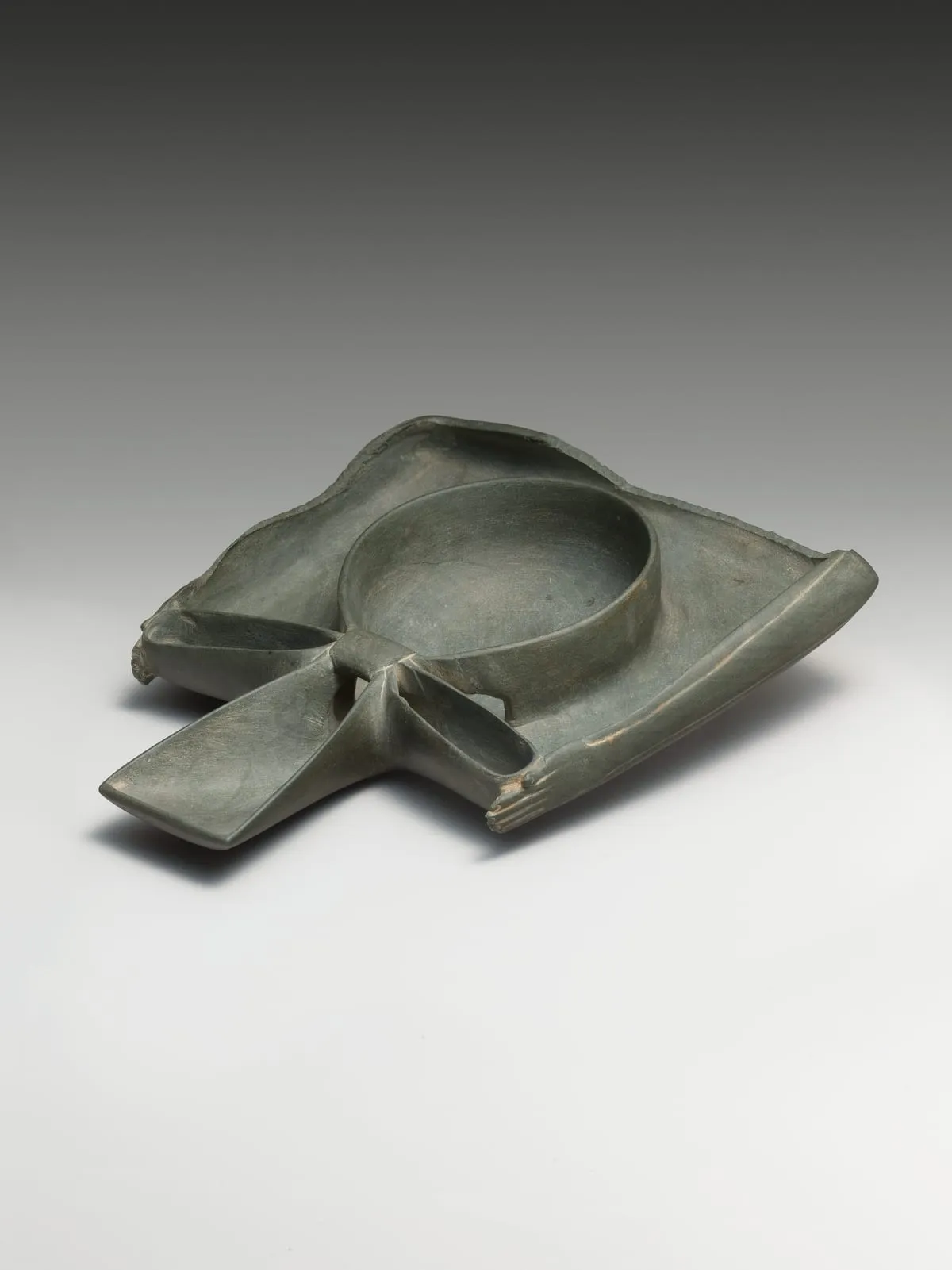 Flache Schale aus grüngrauem Stein, schräg von oben aufgenommen. In der Mitte liegt eine runde Vertiefung. Aus ihr wachsen nach zwei Seiten flache, langgestreckte Fortsätze, die in ausgearbeiteten Händen mit erkennbaren Fingern enden — die Schale hat die Form zweier erhobener Arme. Die Ränder sind unregelmäßig und teils bestoßen.