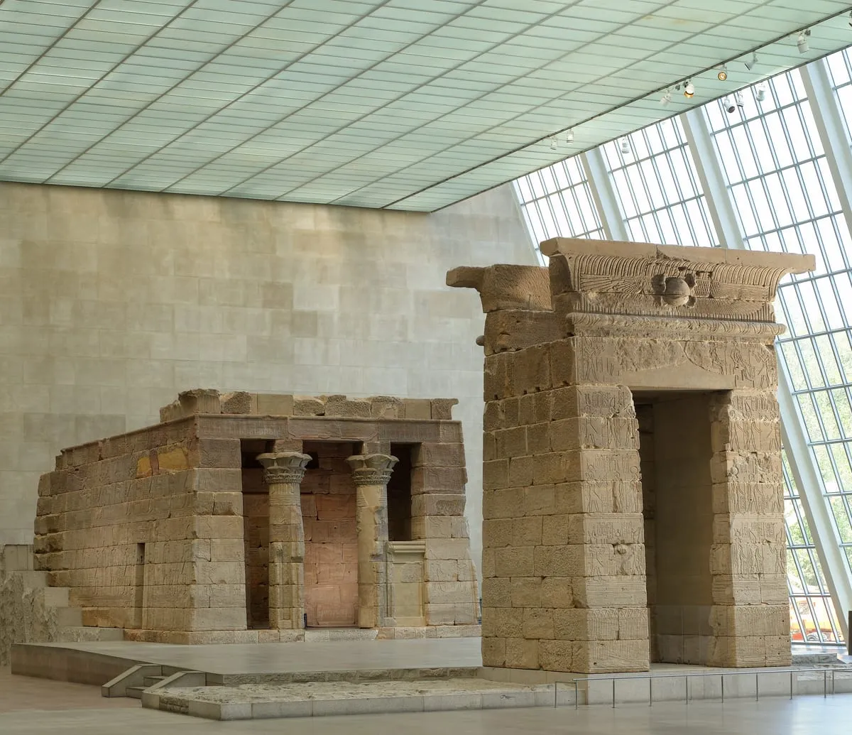 Ein antiker Sandsteinbau in einer modernen Museumshalle. Links steht ein niedriges rechteckiges Gebäude mit erhaltenen Außenwänden; die Front öffnet sich zwischen zwei Säulen mit Pflanzenkapitellen auf einen rötlich getönten Innenraum. Rechts davor steht frei ein wesentlich höheres Torbauwerk mit breiter Durchgangsöffnung, abgeschlossen von einer Hohlkehle mit geflügelter Sonnenscheibe. Beide Bauten sind über und über mit flachen Reliefs und Hieroglyphenkolumnen bedeckt und stehen auf einem hellen Steinpodest. Den Hintergrund bilden eine glatte helle Wand und eine schräg ansteigende, engmaschig gerasterte Glasfassade; von der Decke hängen Strahler an Schienen.