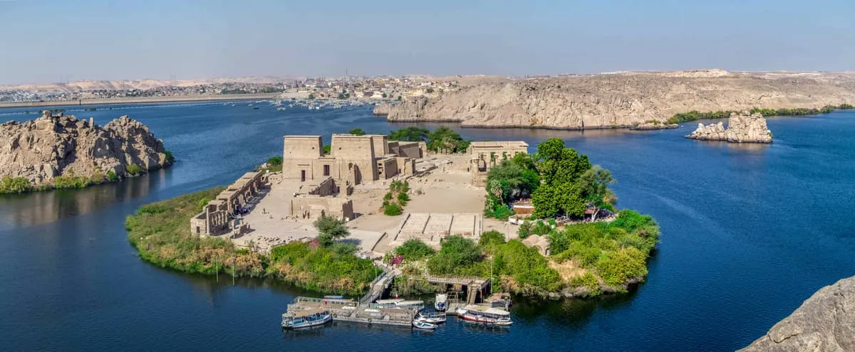 Weiter Blick von oben auf eine kleine, vollständig von tiefblauem Wasser umgebene Insel. Auf ihrer Mitte steht eine dichte Gruppe niedriger, ockerfarbener Steinbauten mit glatten Wänden und flachen Dächern; davor liegt eine gepflasterte Freifläche mit einer breiten Treppe zum Ufer. Der Inselrand ist von grünem Buschwerk und Bäumen gesäumt, am unteren Bildrand liegen mehrere weiße Ausflugsboote an einem Steg. Ringsum ragen kahle, hellbraune Felsklippen aus dem Wasser; im Hintergrund zieht sich eine Stadt am Ufer entlang, darüber ein dunstiger, heller Himmel.