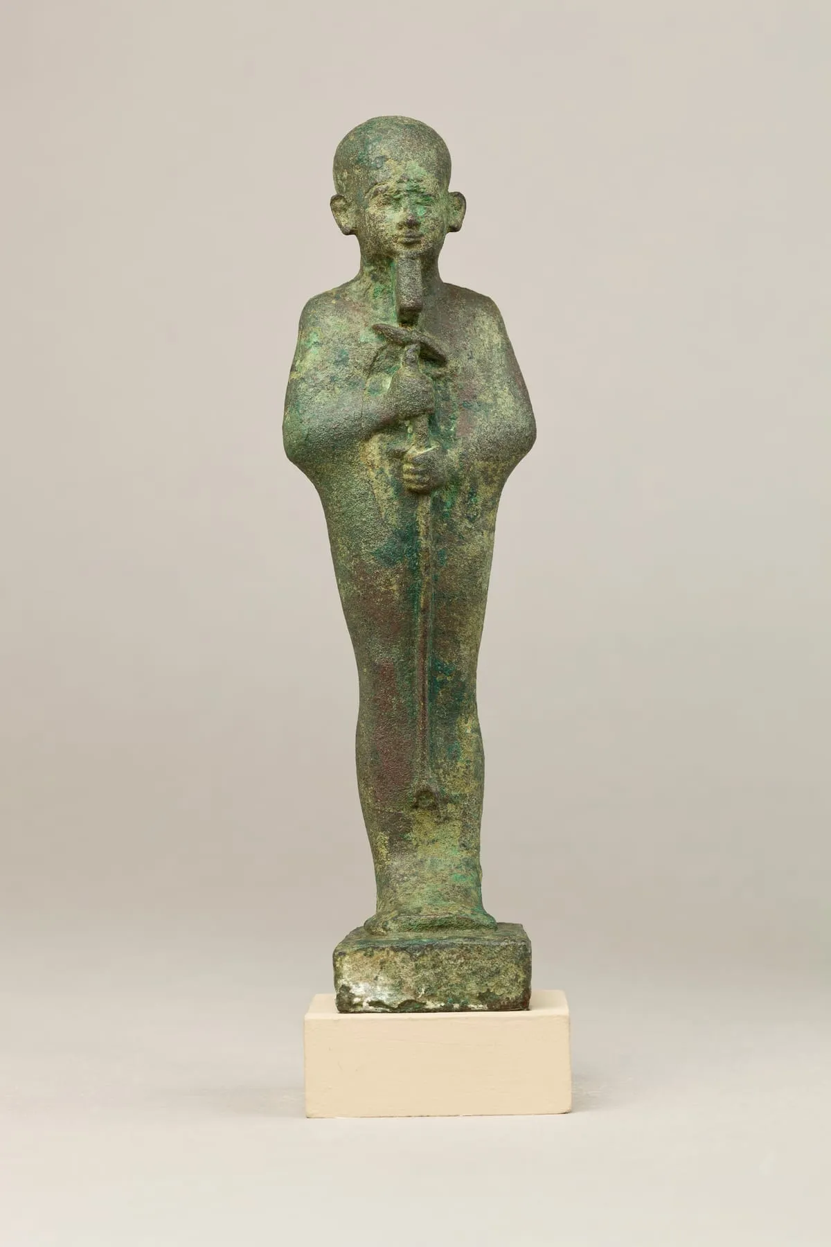 Stehende Bronzefigur in Mumienform auf einer angearbeiteten Standplatte, grün und rötlich patiniert. Der Kopf trägt eine glatte, eng anliegende Kappe; die Ohren stehen frei, das Gesicht ist fein modelliert, vom Kinn hängt ein kurzer gerader Bart. Der Körper ist glatt umwickelt. Beide Hände treten vorn aus der Umwicklung, eine über der anderen, und umfassen einen langen senkrechten Stab, der bis zur Standplatte reicht.