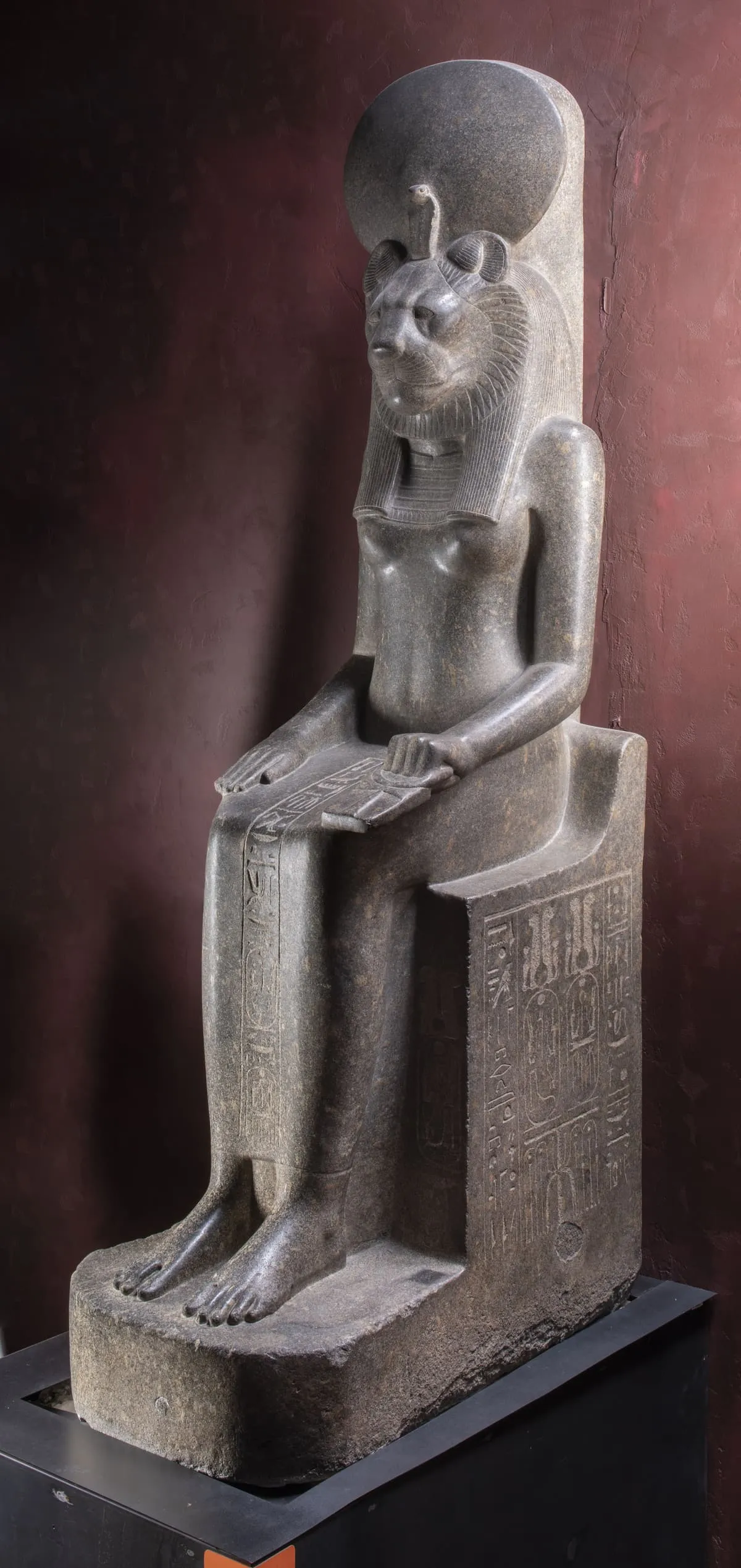 Sitzende Steinfigur, schräg von vorn aufgenommen. Der Kopf ist der einer Löwin mit gerundeter Schnauze und kleinen, nach vorn gedrehten Ohren; dahinter steht eine große runde Scheibe auf, an deren Vorderseite sich eine kleine Schlange aufrichtet. Vom Kopf fällt eine fein gesträhnte Perücke in zwei Strängen über die Schultern. Der Körper ist der einer schlanken Frau; die Hände liegen flach auf den Oberschenkeln, eine davon umfasst einen langgestreckten Gegenstand, der quer über dem Schoß liegt. An der Seitenwange des Sitzes und über den Beinen stehen eingetiefte Hieroglyphen in Kolumnen. Die Füße stehen nebeneinander auf einer Bodenplatte, die Zehen sind einzeln ausgearbeitet. Der Stein ist dunkel und feinkörnig; der Hintergrund ist eine dunkelrote Wand.