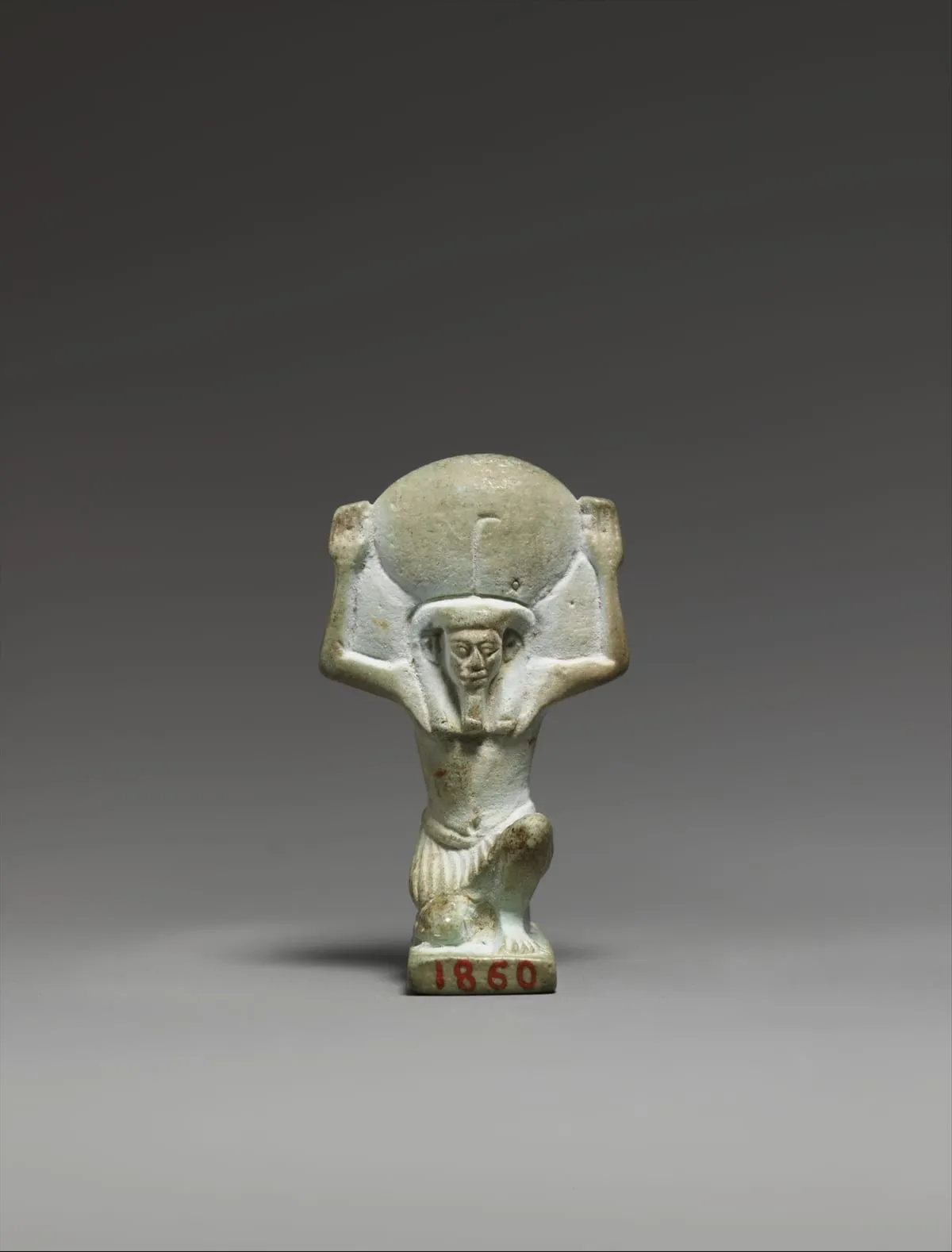 Kleine Amulettfigur aus hellgrüner Fayence. Ein Mann kniet, beide Arme sind über den Kopf erhoben und stützen mit den Händen eine große runde Scheibe. Er trägt einen kurzen gefältelten Schurz; Gesicht und Perücke sind flach ausgearbeitet. Auf der Standplatte steht in roter Farbe eine Nummer.