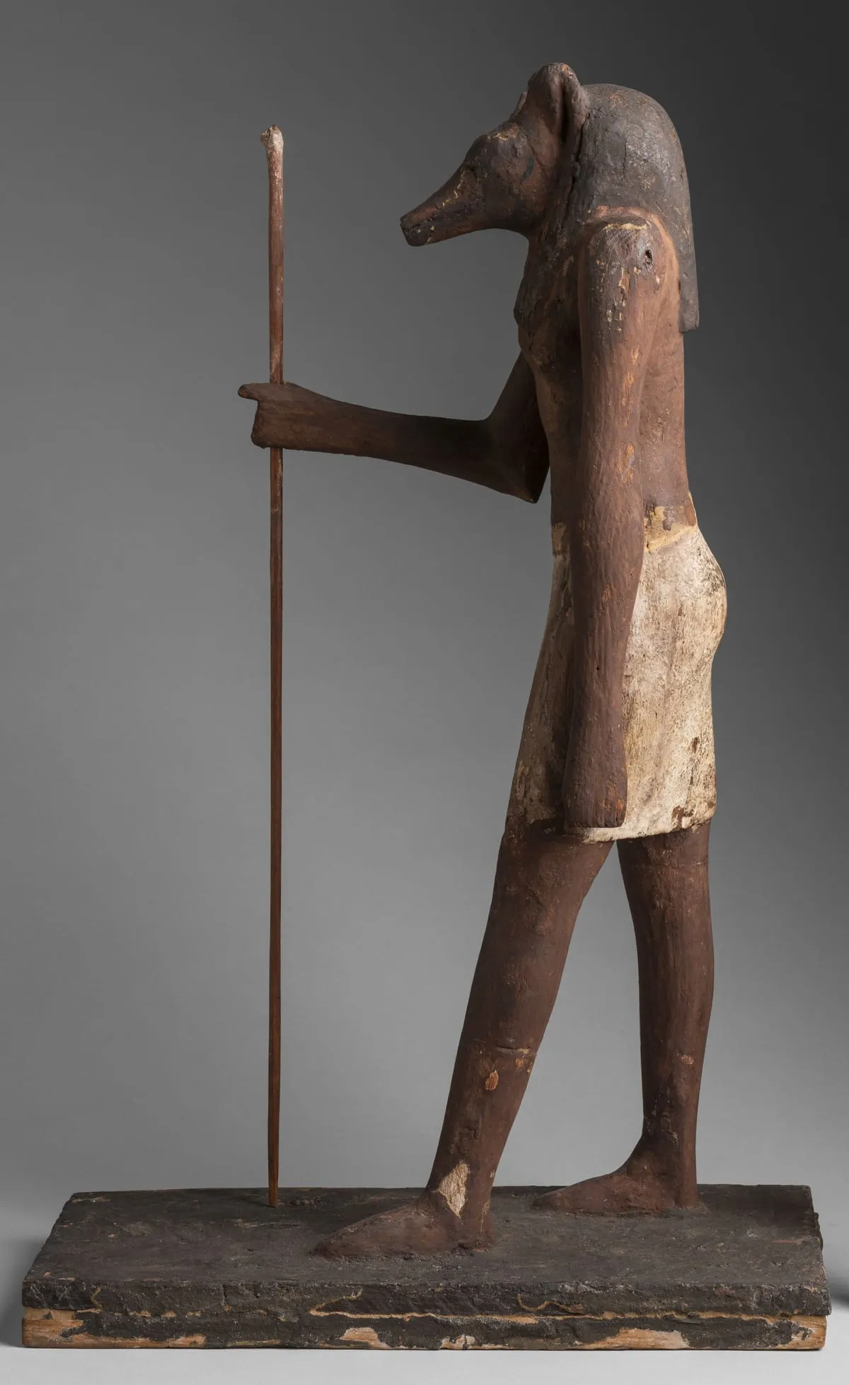 Stehende Holzfigur im Profil nach links, auf einer rechteckigen Holzplatte. Der Körper ist der eines schreitenden Mannes mit nacktem Oberkörper und weißem, knielangem Schurz; das Holz ist rotbraun und an vielen Stellen abgerieben. Der Kopf ist der eines Tieres mit langer, leicht abwärts gebogener Schnauze und stumpf abgesetzter Spitze; darüber steht ein hohes Ohr auf. Vom Kopf fällt eine dunkle Perücke auf die Schulter. Ein Arm ist nach vorn gestreckt, die Hand umfasst einen dünnen, hoch aufragenden Stab; der andere hängt am Körper herab.