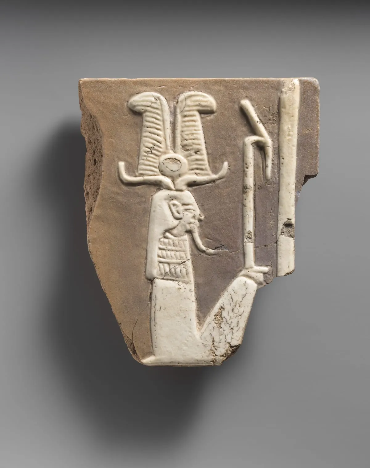 Bruchstück einer Fayencekachel: auf braunviolettem Grund hebt sich eine Figur in weißem Relief ab. Ein Mann sitzt im Profil nach links, das Knie angezogen, und trägt einen langen Kinnbart und einen breiten Halskragen. Auf dem Kopf sitzt eine hohe Krone aus zwei gerippten Federn mit einer Scheibe zwischen ihnen; darunter schwingen zwei geriffelte Widderhörner waagerecht zu den Seiten aus. Rechts steht ein langes Zepter mit gegabeltem Fuß.