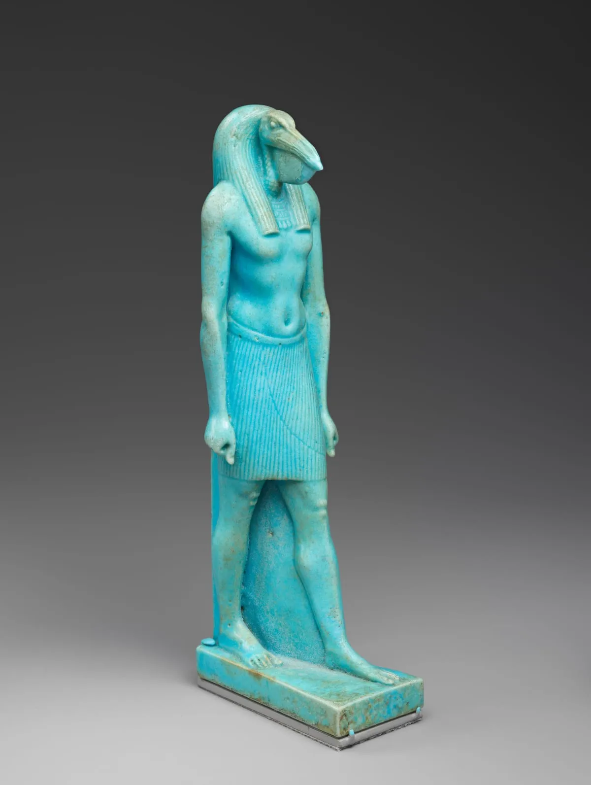Stehende Figur aus leuchtend türkiser Fayence auf einer flachen Platte. Der Körper ist der eines schreitenden Mannes mit nacktem Oberkörper, breitem Halskragen und kurzem gefältelten Schurz; ein Bein ist vorgesetzt. Der Kopf ist der eines Ibis mit langem, abwärts gebogenem Schnabel. Vom Kopf fällt beidseits eine gesträhnte Perücke auf die Brust.