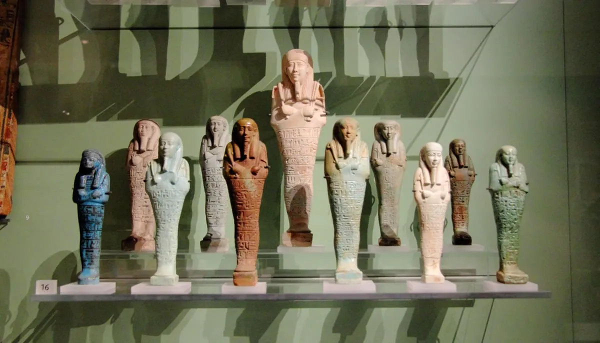 Eine Reihe von elf mumienförmigen Figuren in einer Museumsvitrine, aufgestellt in zwei Ebenen. Jede Figur hat einen glatten, säulenartigen Körper ohne abgesetzte Beine, eine breite Strähnenperücke und über der Brust gekreuzte Arme, die kurze, gebogene Geräte halten. Der Körper ist unterhalb der Arme vollständig mit waagerechten Zeilen kleiner Zeichen bedeckt. Die Figuren sind unterschiedlich gefärbt — kräftig blau, hellgrün, weiß, rotbraun; eine größere in der Mitte ist rosa und trägt einen aufwendiger gearbeiteten Kopfschmuck.