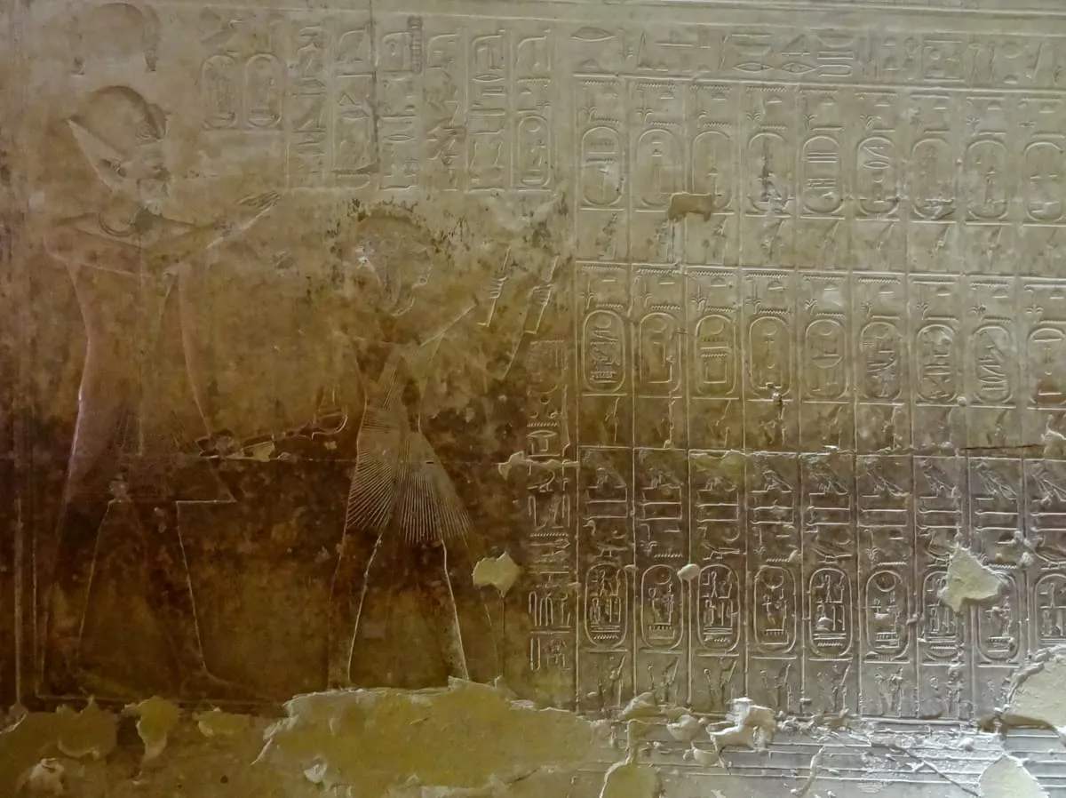 Flachrelief auf heller Kalksteinwand. Links stehen zwei Gestalten im Profil nach rechts, die vordere mit erhobenem Arm; ihre Umrisse sind durch dunkle Verfärbungen teilweise verdeckt. Rechts davon füllt ein Raster den Rest der Wand: drei übereinanderliegende waagerechte Reihen aus jeweils rund zehn ovalen Ringen, jeder mit Zeichen gefüllt. Über und zwischen den Reihen stehen weitere Zeichenkolumnen. Am unteren Rand ist der Putz großflächig ausgebrochen.