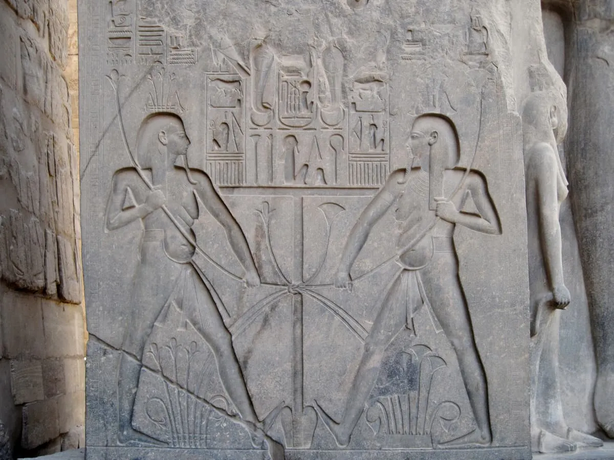 Steinrelief auf einer hochrechteckigen Fläche. Links und rechts kniet je eine männliche Gestalt im Profil, einander zugewandt, mit schulterlanger Perücke, nacktem Oberkörper und kurzem Schurz; auf den Köpfen wachsen Pflanzen. Beide ziehen mit beiden Händen lange Pflanzenstängel zur Mitte, wo sie um einen senkrechten Stab geschlungen und verknotet sind. Über dem Stab stehen zwei Namensringe mit Zeichen. Am unteren Bildrand wachsen dichte Pflanzenbüschel.